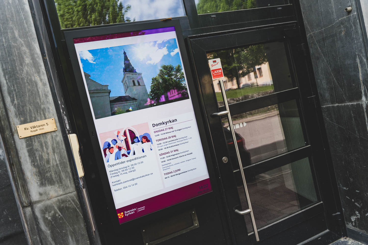 Vertikal digital skärm bredvid entrédörren till Karlstads pastorats expedition, med en bild på Domkyrkan, öppettider och kommande evenemang.