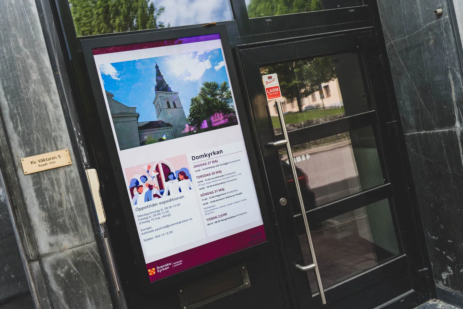 Vertikal digital skärm bredvid entrédörren till Karlstads pastorats expedition, med en bild på Domkyrkan, öppettider och kommande evenemang.