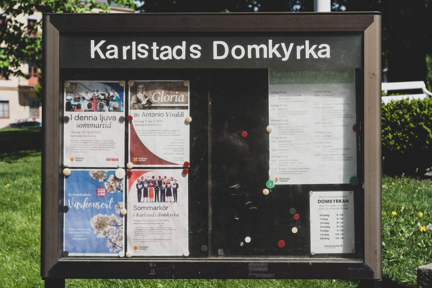 Den gamla anslagstavlan utanför Karlstads Domkyrka, fotograferad rakt framifrån, med påklistrade bokstäver i rubriken och affischer för konserter, körer och gudstjänster.