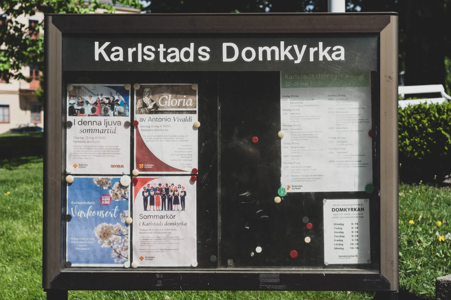 Den gamla anslagstavlan utanför Karlstads Domkyrka, fotograferad rakt framifrån, med påklistrade bokstäver i rubriken och affischer för konserter, körer och gudstjänster.