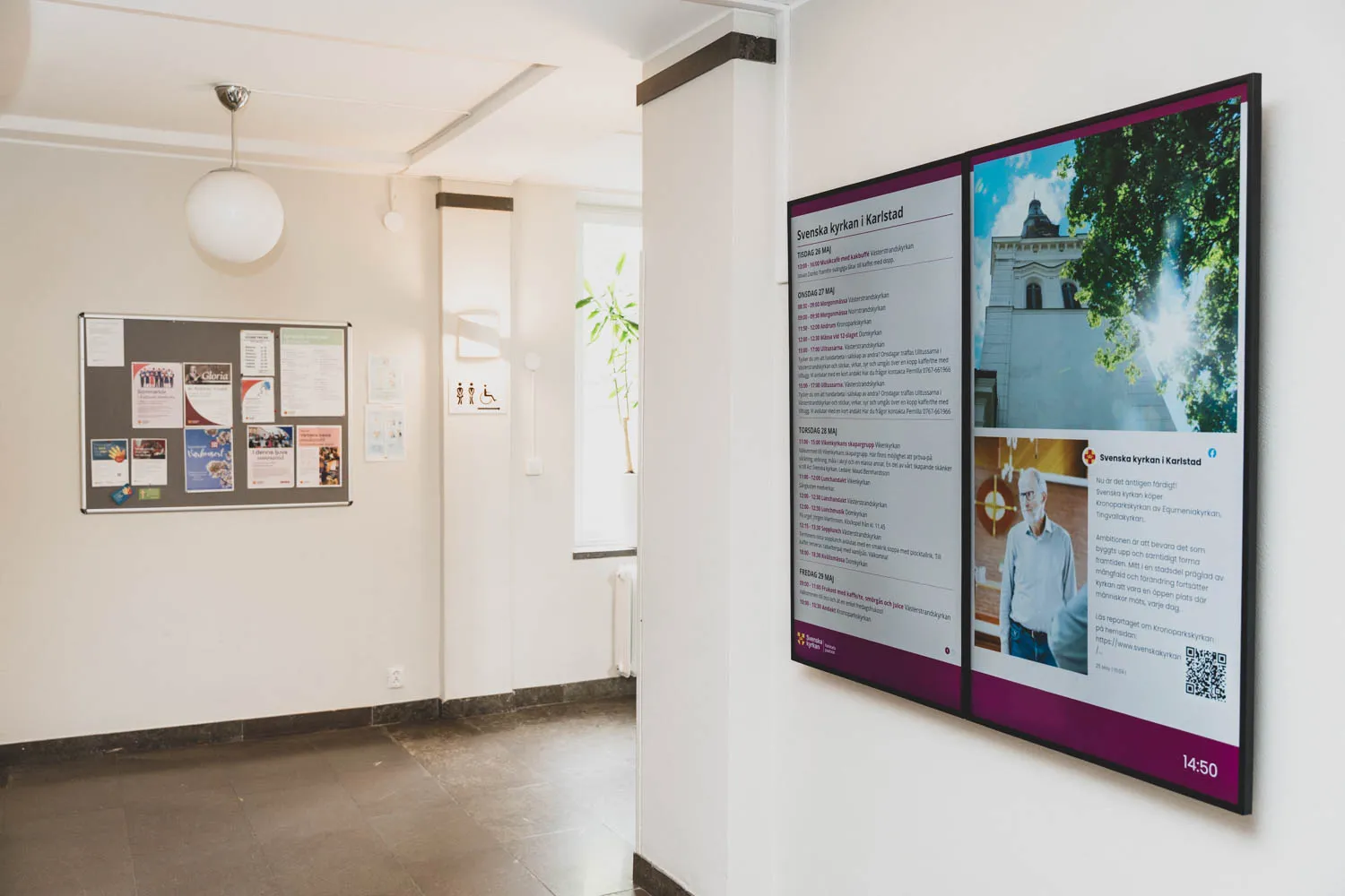 Två digitala skärmar i receptionen hos Karlstads pastorat som visar veckans program och bilder, med den gamla anslagstavlan synlig i bakgrunden.